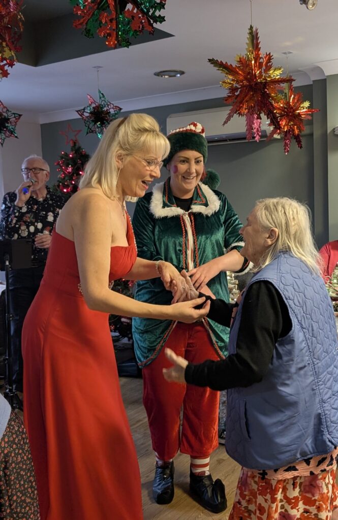 A festive scene where an older woman is smiling and holding hands with two women - one dressed in a red gown and the other in an elf costume. They are dancing together in a room decorated with colourful hanging ornaments and Christmas decorations, while other people enjoy the celebration in the background.