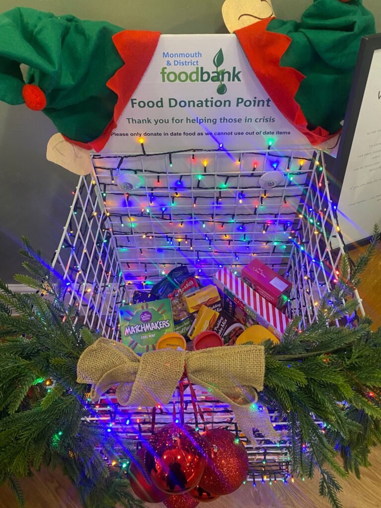 A festive food donation basket for Monmouth & District Foodbank, decorated with Christmas lights, greenery, bows, and large red baubles. A sign at the top reads 'Food Donation Point – Thank you for helping those in crisis.' Inside the basket are donated food items such as chocolate, biscuits, and packaged goods.
